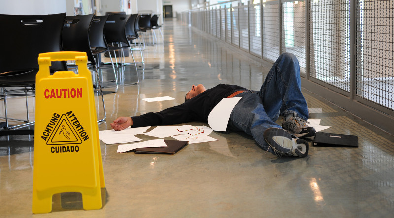 Hình ảnh người đàn ông nằm ngã trên sàn  nhà với biển cảnh báo trượt ngã màu vàng gần vị trí xảy ra tai nạn